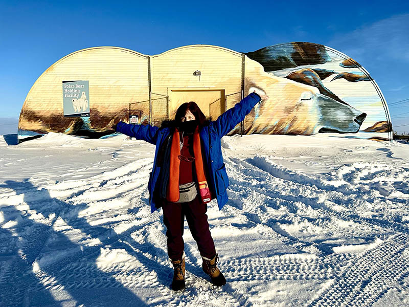 Rozanne, in orange scarf, stands in front of polar bear jail in Churchill.